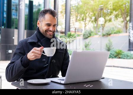 Lächelnder Geschäftsmann im schwarzen Mantel, der eine Tasse Tee trinkt und über einen Laptop mit einem Videoanruf spricht, während er tagsüber am Tisch im Café sitzt Stockfoto