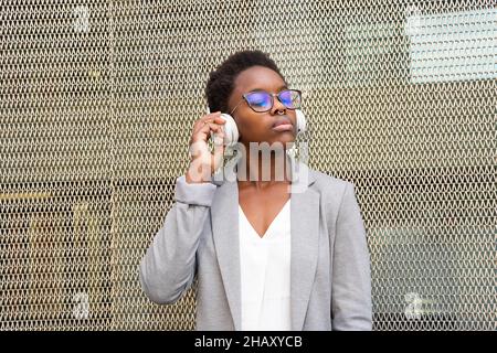 Junge afroamerikanische Frau in elegantem Outfit und Brillen, die Musik in kabellosen Kopfhörern hören, während sie mit geschlossenen Augen steht Stockfoto