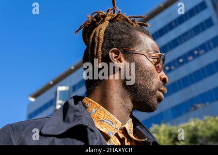 Porträt eines jungen bärtigen afroamerikanischen Mannes in lässigem Hemd und blauem Mantel mit Sonnenbrille und Dreadlocks im Stadtpark an sonnigen Tagen Stockfoto