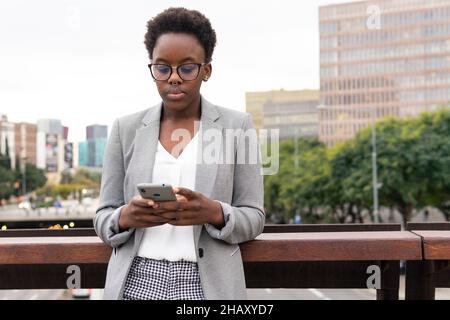 Junge schwarze Angestellte mit Brille und formeller Jacke, die auf dem Balkon des Business Centers auf dem Mobiltelefon herumsteht Stockfoto