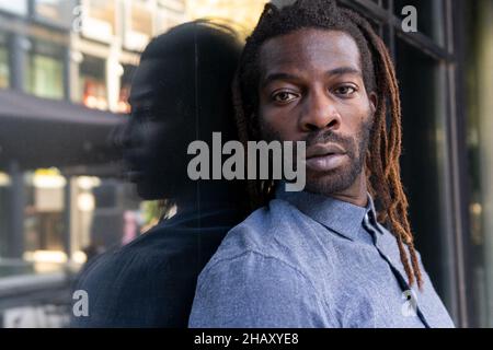 Selbstbewusster afroamerikanischer Mann in legerem Denim-Hemd mit den Armen, die tagsüber in der Nähe des Gebäudefensters gefaltet sind und die Kamera betrachten Stockfoto