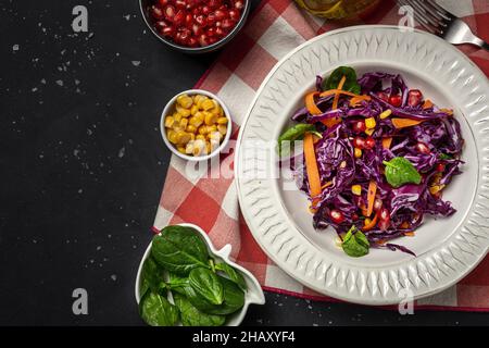 Hausgemachter purpurner Kohlsalat mit Mais, Karotten, Granatapfel und Spinat auf dunklem Grund. Veganes Lebensmittelkonzept. Gesunde Ernährung Stockfoto