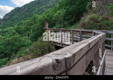 Seitenansicht einer nicht erkennbaren weiblichen Reisenden, die auf einer hölzernen Fußgängerbrücke über den Fluss Mao in Galicia Sacra in Spanien steht Stockfoto