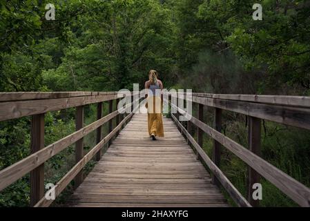 Rückansicht einer unkenntlich Reisenden Frau, die auf einer hölzernen Fußgängerbrücke über den Fluss Mao in Galicia Sacra in Spanien geht Stockfoto