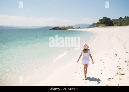 Rückansicht des Ganzkörpers fröhliche Hündin im Sommer-Outfit und Hut, die am weißen Sandstrand der Cies-Inseln in Galicien, falls Spanien, entlang des türkisfarbenen Meeres spazieren Stockfoto