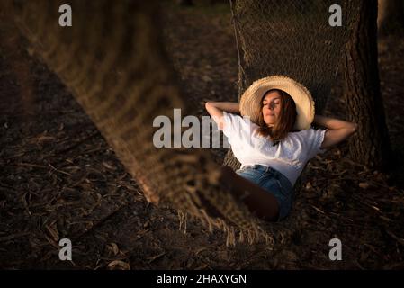 Junge Barfußweibin Strohhut und Jeans-Shorts liegen auf gestrickter Hängematte und entspannen in Cambados Natur in Spanien Stockfoto