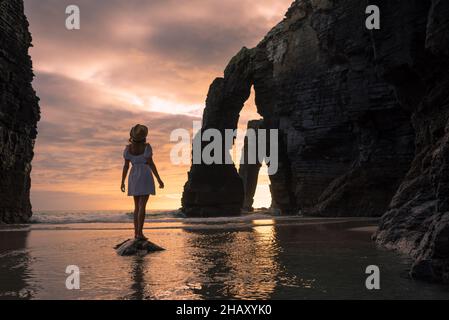 Rückansicht des Weibchens in Kleid, das auf Felsen in seichtem Meerwasser wieder Berge steht und den Sonnenuntergang in Galicien betrachtet Stockfoto