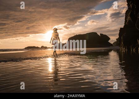 Silhouette einer einsamen, nicht erkennbaren Frau, die am Sandstrand der Kathedralen in der Nähe von felsigen Klippen steht und den malerischen Sonnenuntergang am Himmel bewundert Stockfoto