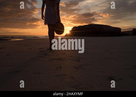 Verkürzte Silhouette einer einsamen, nicht erkennbaren Frau, die am Sandstrand der Kathedralen in der Nähe von felsigen Klippen steht und den malerischen Sonnenuntergang am Himmel bewundert Stockfoto