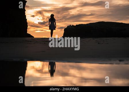 Silhouette einer einsamen, nicht erkennbaren Frau, die am Sandstrand der Kathedralen in der Nähe von felsigen Klippen steht und den malerischen Sonnenuntergang am Himmel bewundert Stockfoto