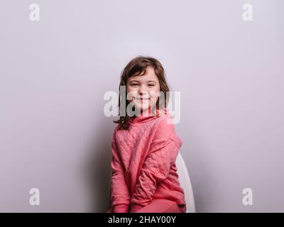 Nettes glückliches Mädchen in rosa Kleidung lächelnd und Blick auf die Kamera, während auf dem Stuhl gegen die graue Wand sitzen Stockfoto