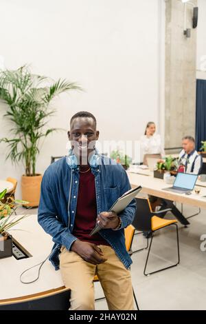Optimistischer afroamerikanischer Mann mit Kopfhörern am Holzarbeitsplatz mit Mitarbeitern am Tisch mit Laptops Stockfoto