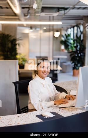 Lächelnde Managerin in eleganter Kleidung, die am Computer tippt, während sie während der Arbeit im modernen hellen Büro am Schreibtisch sitzt Stockfoto