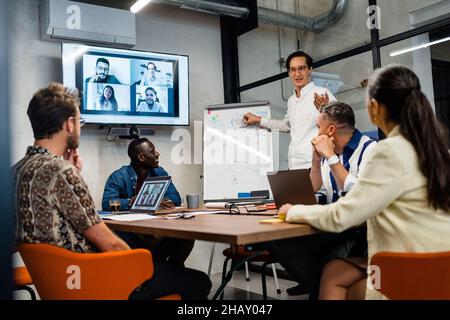 Mann, der während eines Online-Meetings in der Nähe von Mitarbeitern im Konferenzraum ein Geschäftsprojekt am Whiteboard vorführt Stockfoto
