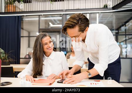 Positive Frau in formeller Kleidung, die Kollegen beim Surfen am Tisch mit dem Veranstalter anschaut, während sie in einem hellen, modernen Büro zusammenarbeitet Stockfoto
