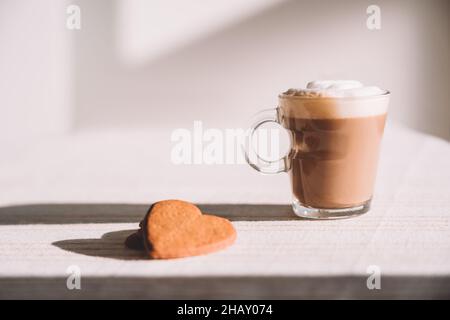 Ein Glas schaumigen Latte Macchiato wird am sonnigen Tag in der Küche mit herzförmigen Plätzchen auf dem Tisch serviert Stockfoto