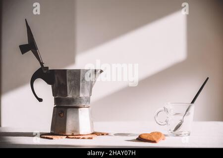 Mokakatopf aus Metall mit geöffnetem Deckel, der an sonnigen Tagen auf einem weißen Tisch in der Nähe von Glastassen und herzförmigen Keksen aufgestellt wird Stockfoto