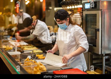 Durch ein Glas weiblicher Köchin mit weißer Jacke, die Papierblätter überprüft, während sie an der Theke in der Küche steht und während der Kochzeit fleißige Köchinnen das Essen zubereiten Stockfoto