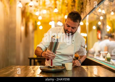 Bärtiger Profi-Koch in weißer Uniform mit Spender, der eine Portion Seeigel in einem feinen Gericht-Restaurant mit Creme ergänzt Stockfoto