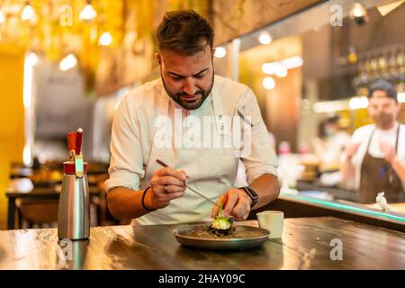 Bärtiger, professioneller Chefkoch, der Zutaten für Seeigel auf dem Teller am Tisch im modernen Restaurant zubereitet Stockfoto