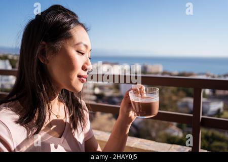 Glückliche asiatische Frau mit einem Glas heißem Getränk in der Hand, während sie auf dem Balkon sitzt Stockfoto
