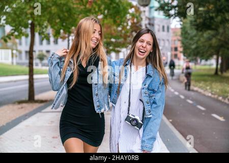 Positive Freundinnen in stilvoller Kleidung umarmten sich beim Spaziergang auf einem gepflasterten Gehweg in der Nähe von Bäumen auf der Straße der Stadt Stockfoto