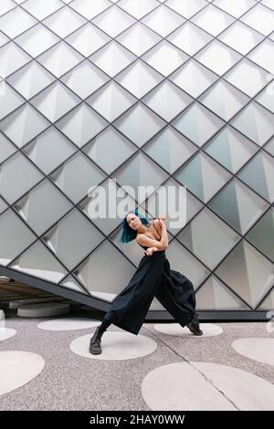 Junge Frau mit blauen Haaren in schwarzem Outfit, die in der Nähe eines modernen Gebäudes auf der Straße der Stadt zeitgenössischen Tanz aufführt Stockfoto