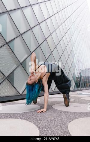 Junge Frau mit blauen Haaren in schwarzem Outfit, die in der Nähe eines modernen Gebäudes auf der Straße der Stadt zeitgenössischen Tanz aufführt Stockfoto