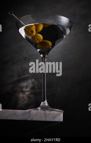 Glas gefüllt mit Alkohol martini mit reifen Oliven auf Spieß auf Rand des Marmortisches vor dunklem Hintergrund im Studio platziert Stockfoto