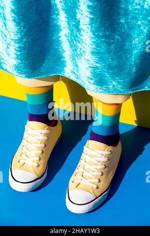 Crop anonyme homosexuelle Person trägt gelbe Turnschuhe und helle Socken mit Regenbogenfarben stehen auf blauer Oberfläche in hellen Raum Stockfoto