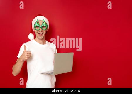 Ein aufgeregter Weihnachtsmann hält Laptop in den Händen und gibt auf rotem Hintergrund die Daumen hoch Stockfoto