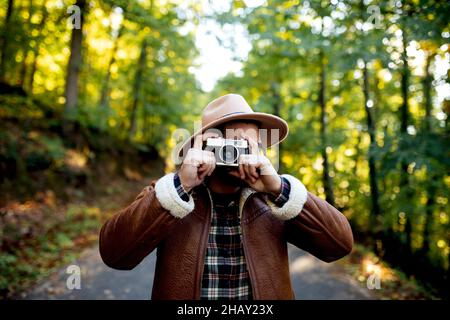 Fotograf in stilvoller Kleidung, der während der Reise auf einem Asphaltweg in der Nähe von hohen grünen Bäumen die Natur fotografiert Stockfoto