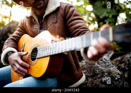 Beschnittene, nicht erkennbare, talentierte bärtige Musikerin in Hut und Oberbekleidung, die auf der Gitarre singt, während sie im Wald neben hohen grünen Bäumen sitzt Stockfoto