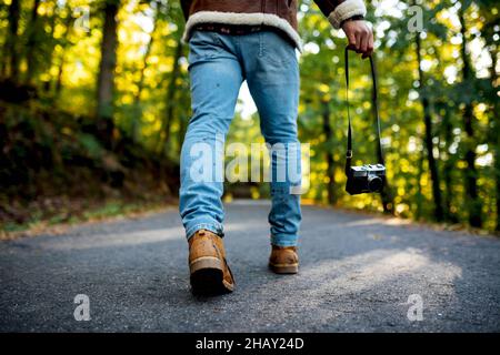 Von unten Rückansicht des zugeschnittenen, unkenntlich coolen Hipster-Typen in trendigem Outfit und Hut, der mit Fotokamera auf einem Asphaltweg in der Nähe von hohen Bäumen läuft Stockfoto
