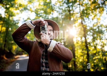 Fotograf in stilvoller Kleidung, der während der Reise mit einer Vintage-Kamera über die Natur fotografiert, während er auf einem Asphaltweg in der Nähe von hohen grünen Bäumen steht Stockfoto