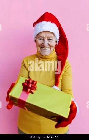Positiv gealtertes Weibchen in rotem Weihnachtsmütze, das während der festlichen Weihnachtsfeiertage auf rosa Hintergrund eine eingewickelte Geschenkbox zeigt Stockfoto