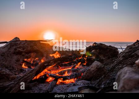 Lagerfeuer mit brennendem Feuerholz während des wunderschönen Sonnenuntergangs am Strand Stockfoto