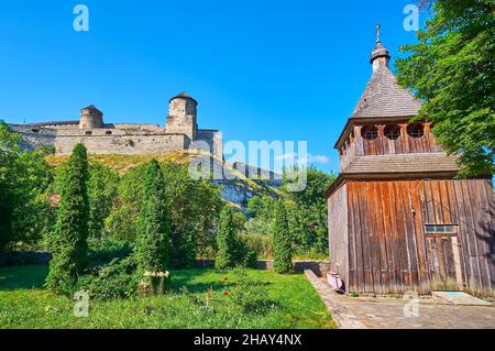 Die hölzerne Kirche Zdvyzhenska (Erhöhung des Heiligen Kreuzes) befindet sich am Fuße des Hügels, umragt mit dem mittelalterlichen Kamianets-Podilskyi Schloss, Ukraine Stockfoto