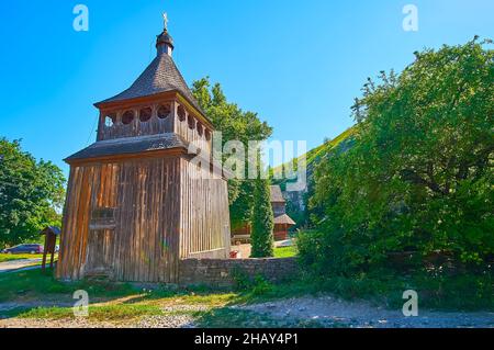 Die ausgebreiteten grünen Bäume rund um den alten hölzernen Glockenturm der historischen Kirche Zdvyzhenska (Erhöhung des Heiligen Kreuzes), in der Schlucht des Smotrych River, Ka Stockfoto