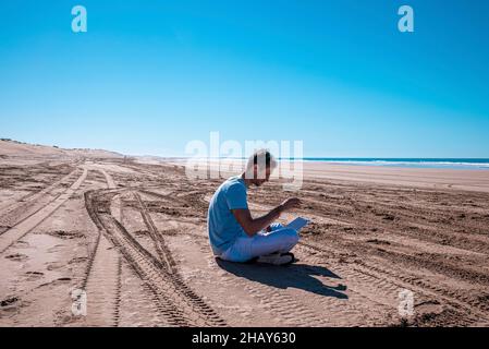 Mann mit Laptop sitzt an einem hellen sonnigen Tag am Strand Stockfoto