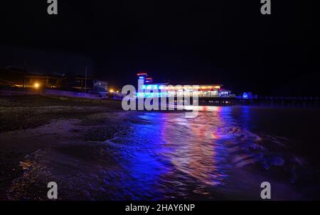 Felixstowe, Suffolk, Großbritannien: Felixstowe Pier bei Nacht beleuchtet mit blauen und gelben Lichtern. Blick auf den Pier vom Strand mit Reflexionen im Meer Stockfoto