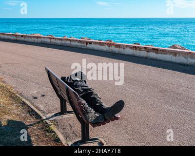 Ein Einzelbettler schläft auf einer Bank an der Mittelmeerküste in Menton, Frankreich Stockfoto