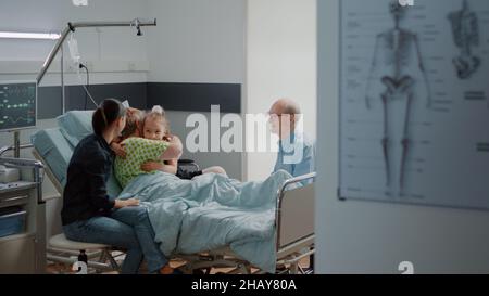 Kind umarmte kranke Patientin im Krankenhausbett beim Familienbesuch mit der Mutter. Nichte besucht eine pensionierte Frau auf der Intensivstation und führt ein Gespräch, um bei der Genesung im Gesundheitswesen zu helfen. Stockfoto
