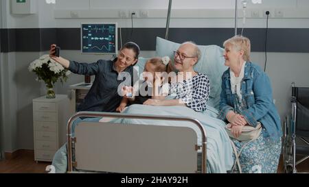 Familie und pensionierter Mann, der beim Besuch im Krankenbett Selfie mit dem Smartphone gemacht hat. Mutter und Kind haben Spaß mit Bildern auf dem Handy, besuchen ältere Patienten mit Krankheit. Stockfoto