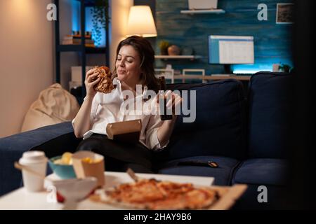 Lächelnde Frau, die auf der Couch sitzt und auf einen leckeren Burger zum Mitnehmen schaut, der eine Bierflasche vor dem Tisch mit Pizza und Junk-Food-Gerichten zum Mitnehmen hält. Fröhliche Person, die leckeren Burger und Lieferservice-Abendessen isst. Stockfoto