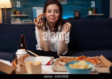 Porträt einer lächelnden Frau nach der Büroarbeit, die ein Stück Pizza mit Heißlieferung isst, während sie am Tisch Fernsehen und ein Schnellimbiss zum Mitnehmen serviert. Fröhliche Person, die auf der Couch sitzt und das Abendessen zum Mitnehmen im fernsehen isst. Stockfoto