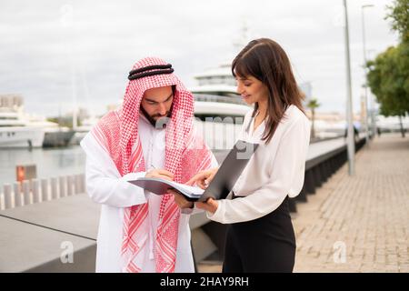 Zufriedener arabischer Mandant, der beim Treffen mit einer Partnerin im Hafen einen Vertrag unterzeichnet, einen Vertrag mit einer lächelnden europäischen Geschäftsfrau geschlossen hat, und ein saudischer Geschäftsmann, der die Unterschrift auf dem Vertragsdokument setzt Stockfoto
