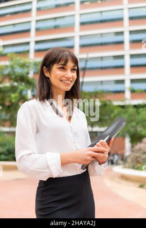 Glückliche attraktive europäische Geschäftsfrau in formeller Kleidung hält Zwischenablage mit Dokumenten und lächelnd, auf der Stadtstraße stehend und wartet auf Kunden oder Geschäftspartner bei der Besprechung Stockfoto