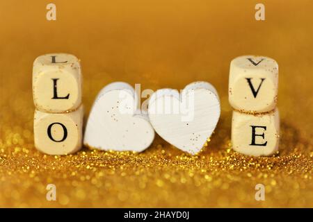 Valentinstag. Liebe und Beziehung. Inschrift Love aus Holzbuchstaben und weißen Herzen auf goldenem Glitzer-Hintergrund mit goldenem Bokeh Stockfoto