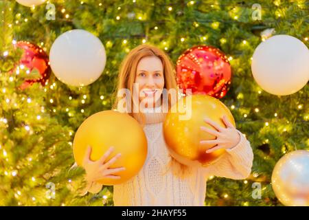 Lächelndes Mädchen hält riesige Weihnachtskugeln auf dem Hintergrund eines eleganten Weihnachtsbaums. Festliches Poster. Herzlichen Glückwunsch Stockfoto